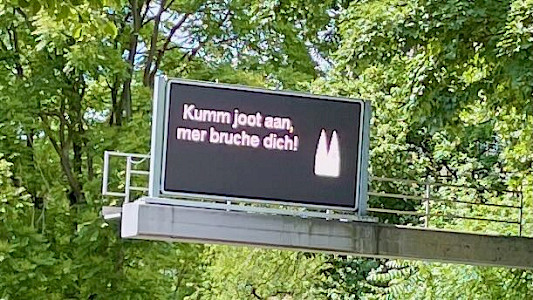 Straßenschild in Köln mit der Aufschrift "Kumm joot aan, mer bruche dich!" (auf Hochdeutsch: „Komm gut an, wir brauchen dich!“)