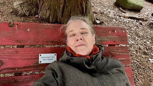 Foto von Sabine mit geschlossenen Augen auf einer roten Liegebank im Wald mit einem Schild "Genießen Sie den Wald!"